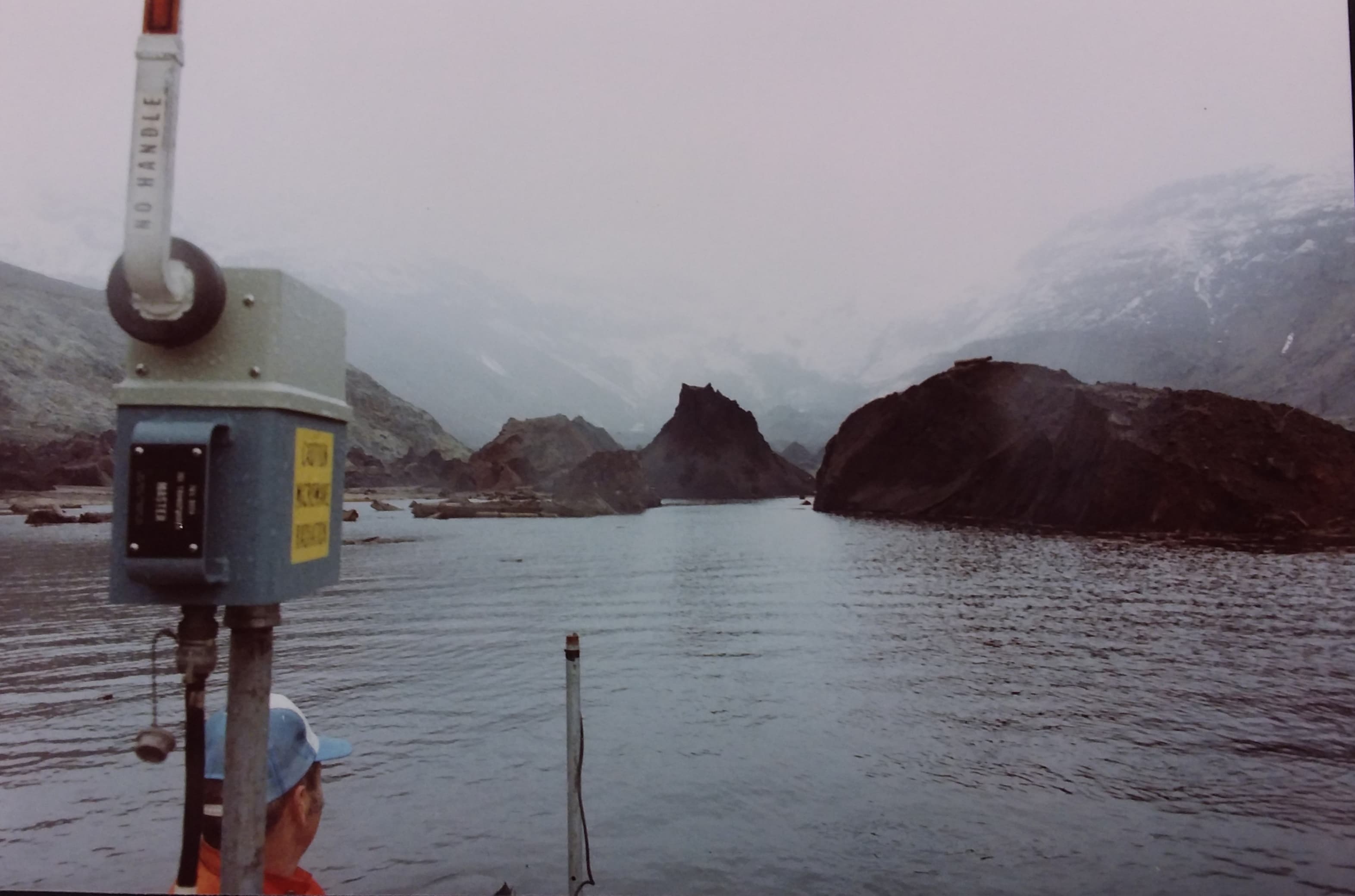 Mid-May 1981  Leonard Nelson in aluminum boat in Bear Cove, West Arm of Spirit Lake with debris avalanche hummocks. Microwave trisponder range finder in boat for use in bathymetric mapping.  USGS K. Tanner